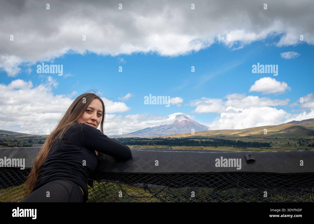 Young woman relaxing and looking at camera with Cotopaxi volcano in the background in the Ecuadorian Andes. Scenic landscape with dramatic clouds. Stock Photo