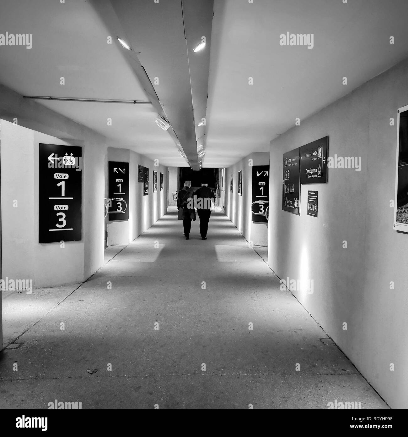 Two people are walking away from the camera in a brightly lit underground tunnel. The tunnel is lined with signs providing directions. France - Smartphone Captured Stock Image