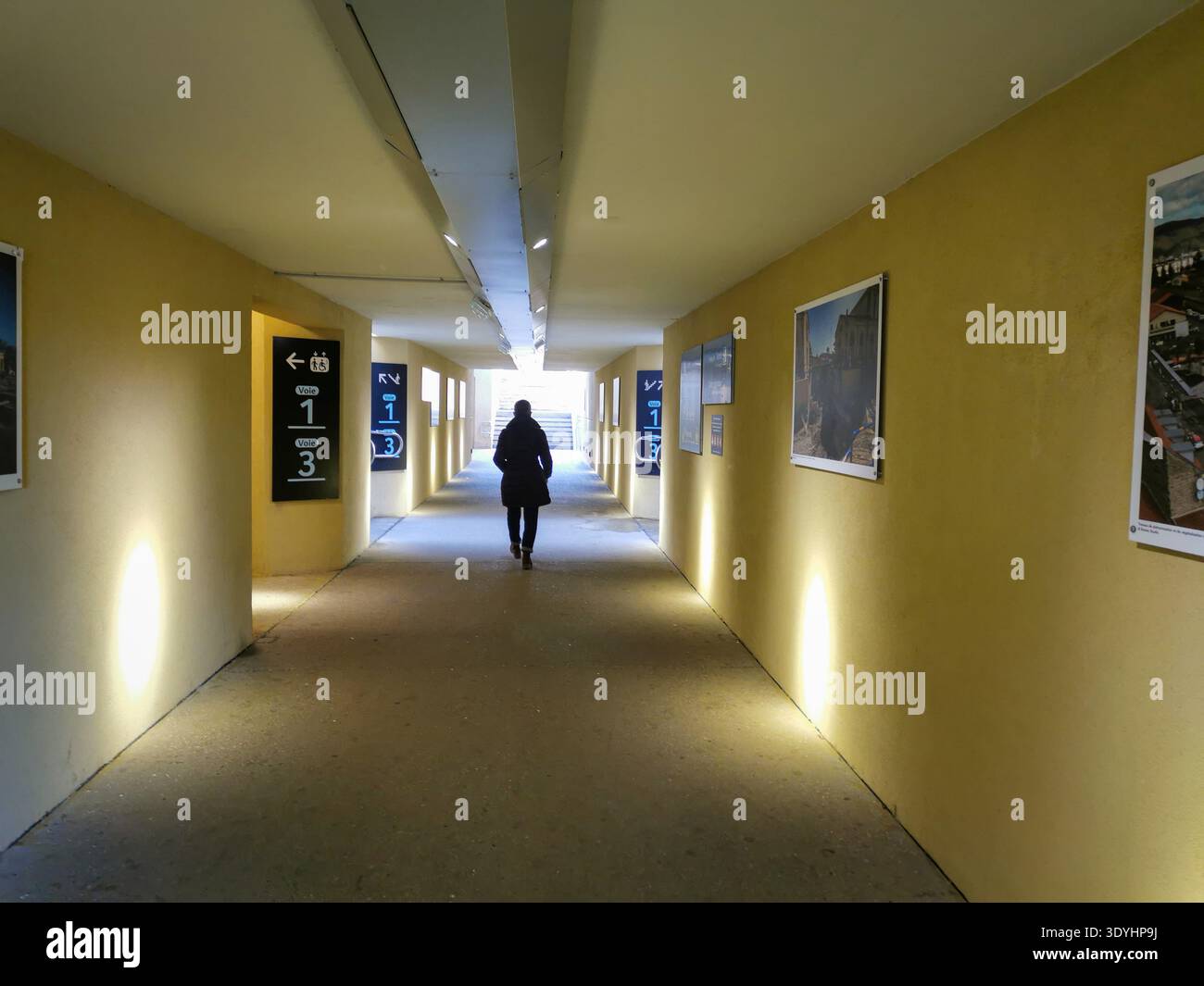 A person walks away from the camera through an underpass. The underpass has yellow walls, signs for train platforms. France - Smartphone Captured Stock Image