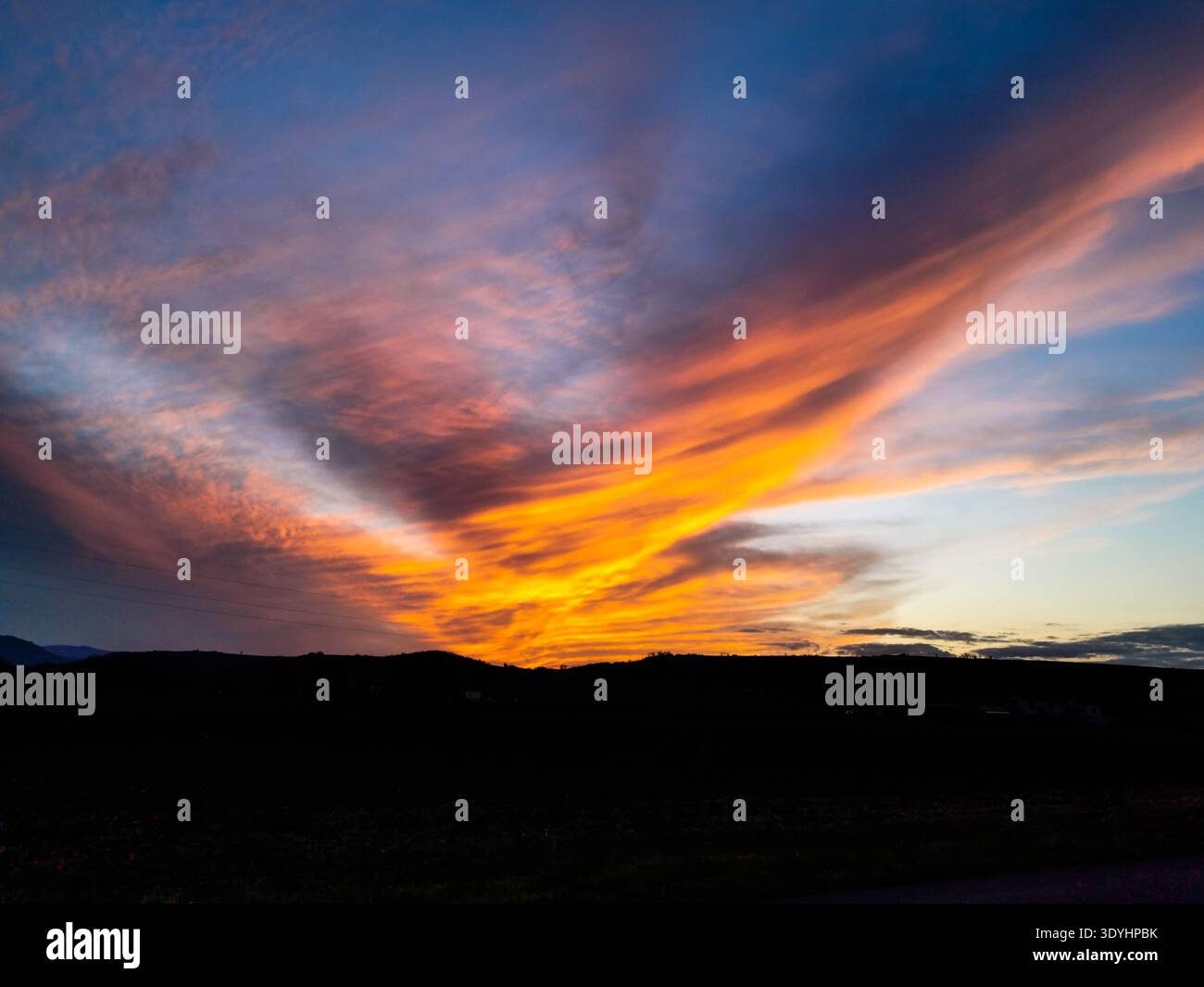 The sun dips below the horizon, creating a spectacular scene where brilliant orange and yellow clouds spread across a deep blue sky. - Smartphone Captured Stock Image