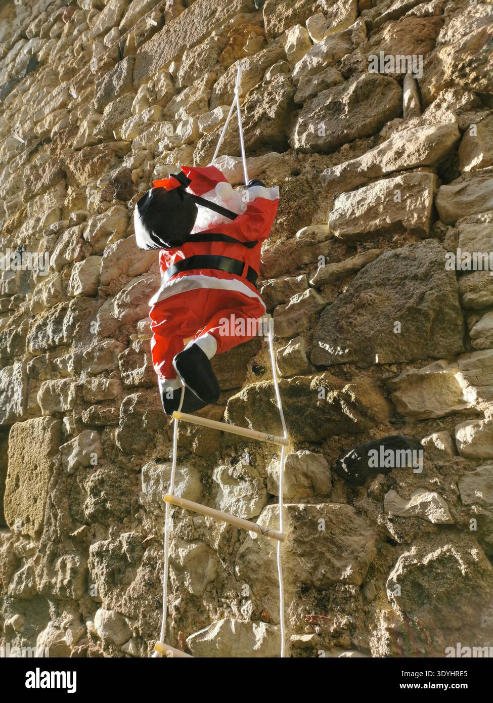 A Santa Claus figure is hanging from a rope ladder and climbing down the side of a rustic stone building. France - Smartphone Captured Stock Image
