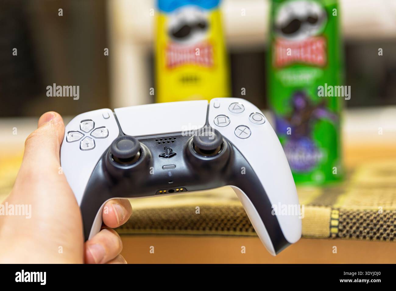 Hand holding a white gaming controller with snacks in the background. Stock Photo