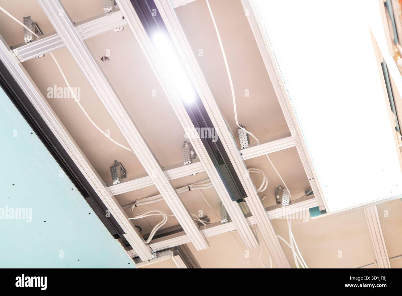 Close-up of a modern suspended ceiling installation featuring LED track lighting, exposed white wiring, and metallic support beams indoors. Stock Photo