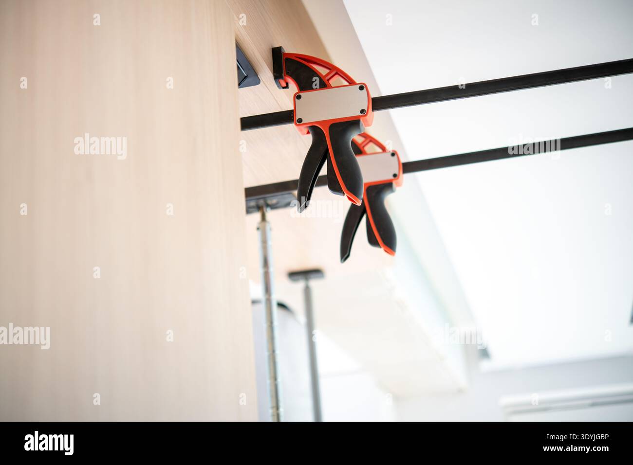 Two red and black pipe clamps securing wooden boards together in a workshop with bright natural lighting and industrial elements. Stock Photo