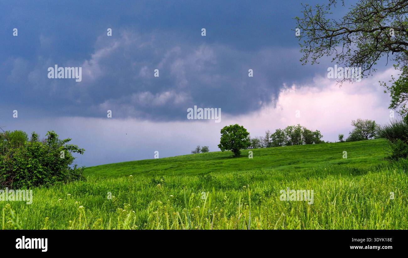 Stormy sky over green hills in Neviano degli Arduini, Italy.A dramatic landscape featuring vibrant green spring meadows under a dark, heavy storm sky. Stock Photo