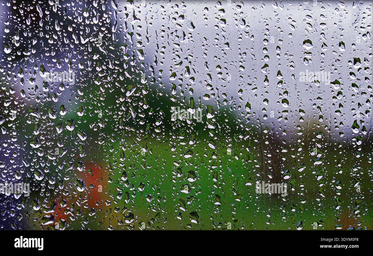 Raindrops cover a window pane. Outside, green trees are partly visible under a cloudy sky. The scene shows a typical rainy day without any people or a Stock Photo