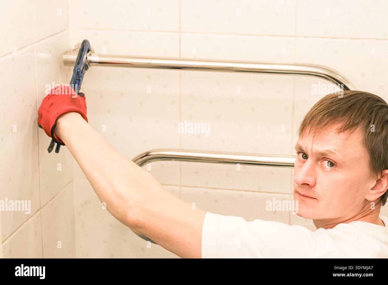 Young man with serious expression in red glove using wrench to adjust chrome rail on white tiled bathroom wall. Stock Photo