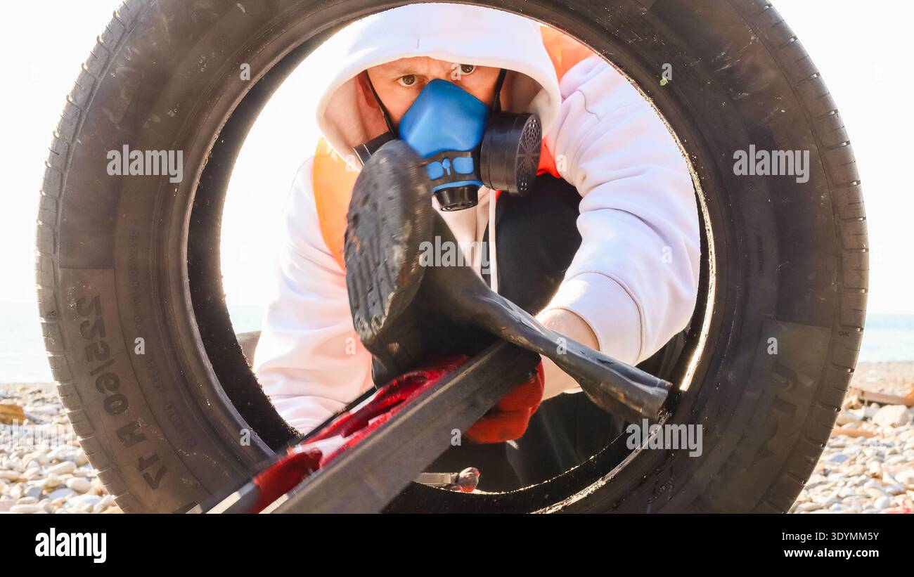 Equipped volunteer uses crowbar to remove debris from old tire on pebble-filled coastline. Stock Photo