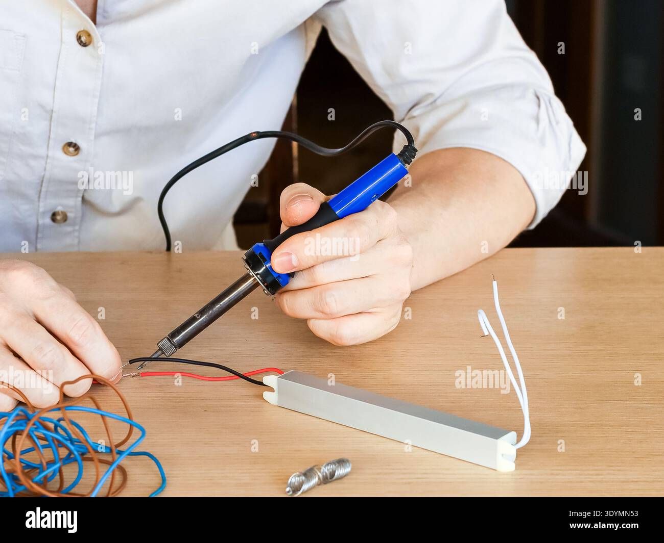 Hands using soldering iron to join red and black wires to silver rectangular module near coiled cables Stock Photo