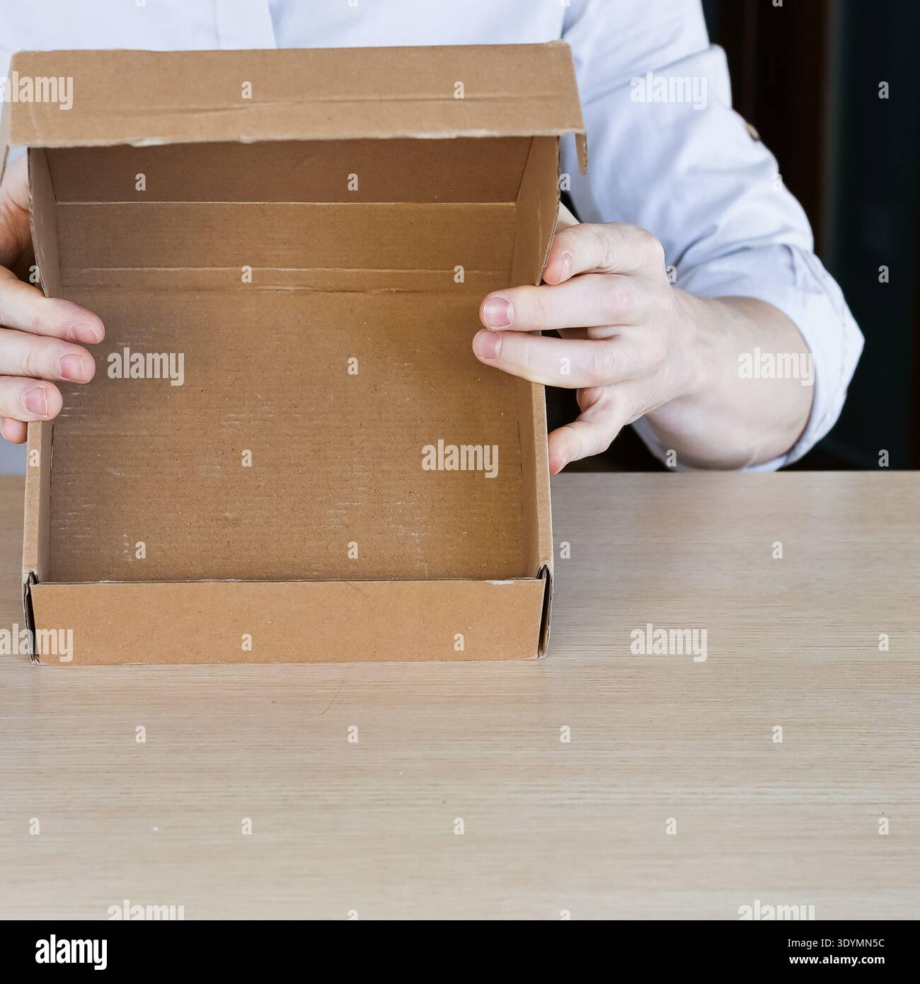 Arms raising small open cardboard box above table, displaying empty interior space. Stock Photo