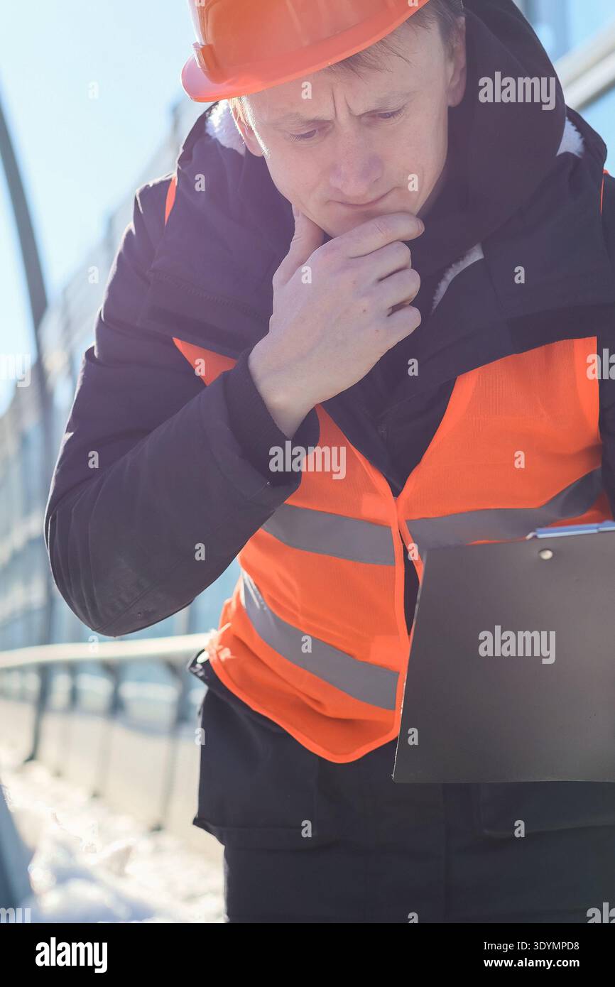 Male engineer in safety gear thoughtfully reviews documents outdoors in snowy urban environment Stock Photo
