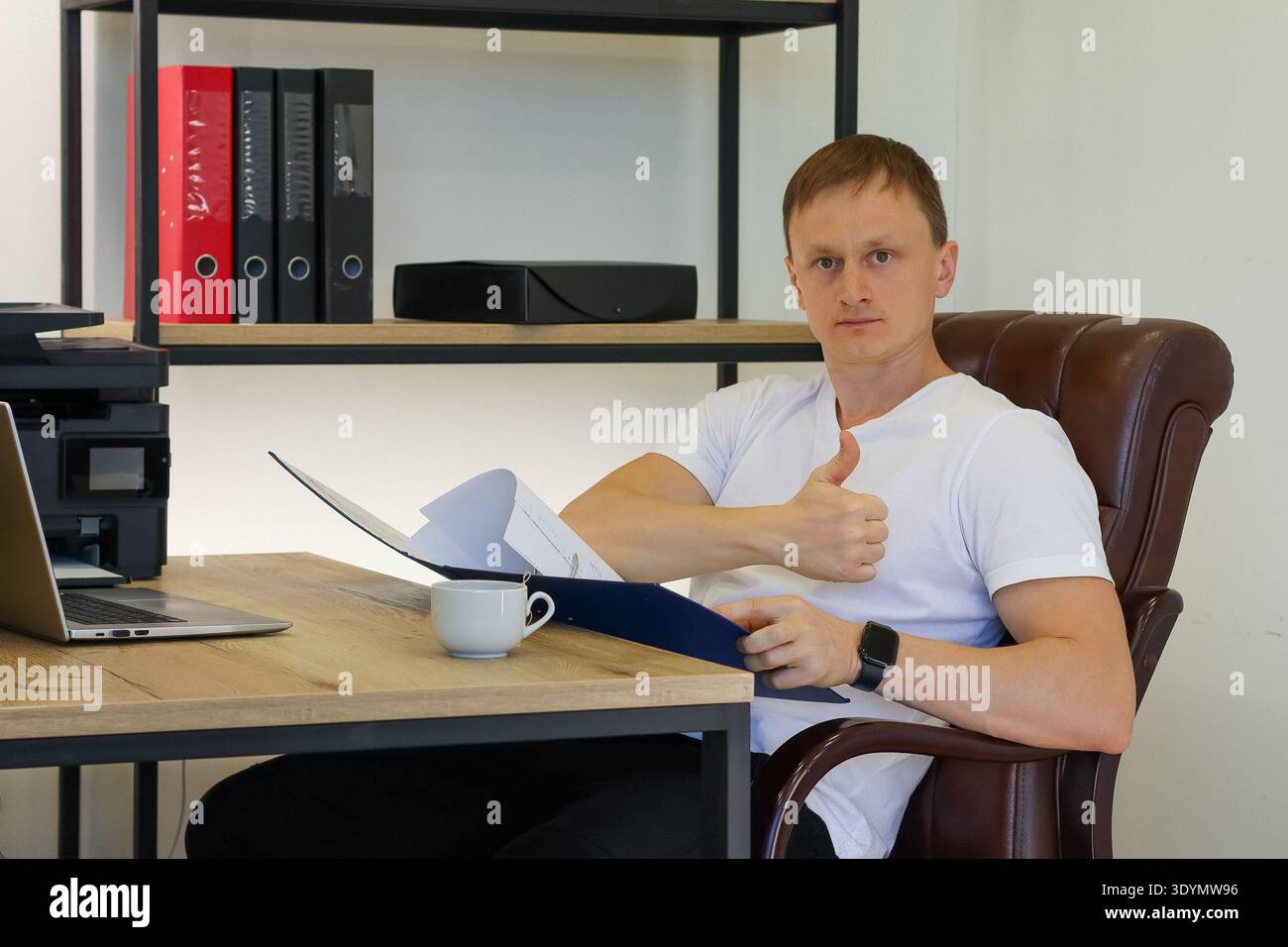 Young professional in office chair gives positive thumb gesture while reviewing blue file contents Stock Photo