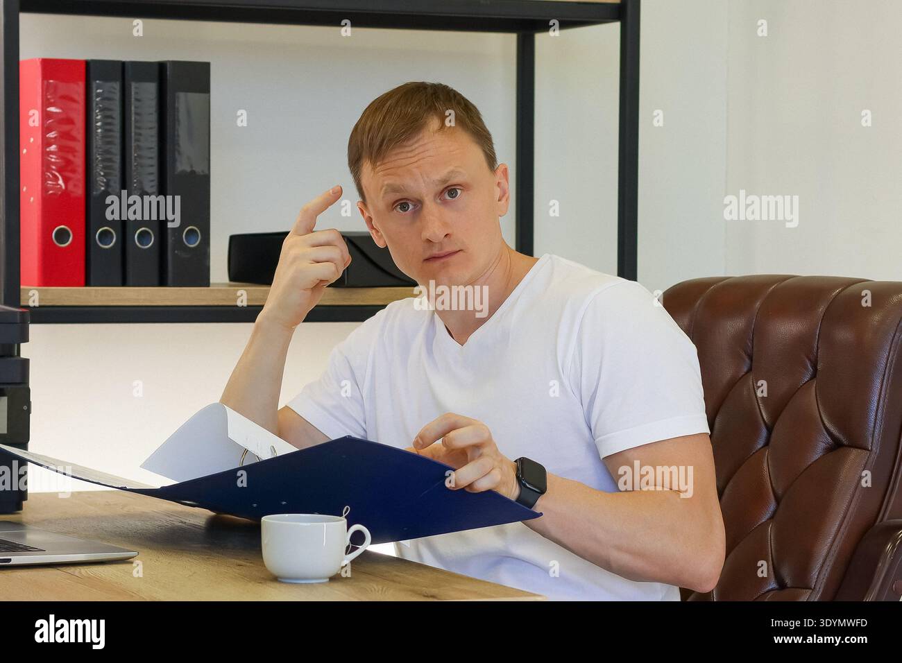 Reflective man tapping finger on temple while holding blue folder with papers at office setup Stock Photo
