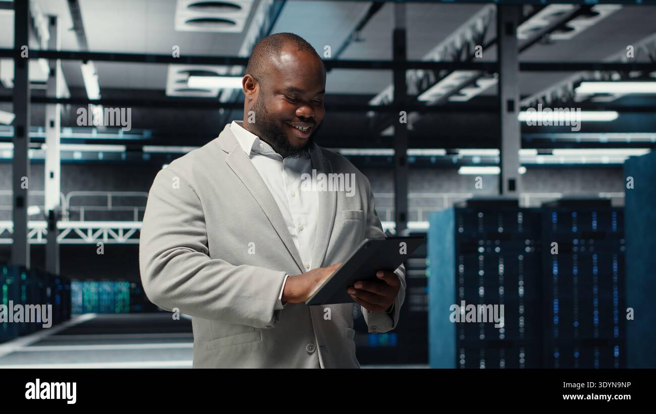 Smiling admin using tablet in in data center maintaining infrastructure for workloads. Joyful IT professional in server farm utilizing high tech server clusters for parallel processing, camera B Stock Photo