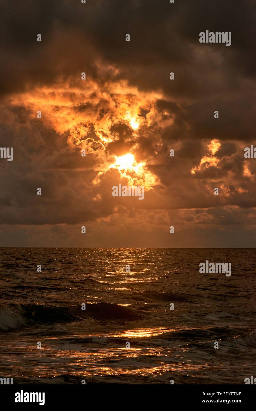 A stunning and dramatic sunset scene over the open ocean, featuring a vibrant, fiery sky with the sun breaking through dark, ominous clouds. The golde Stock Photo