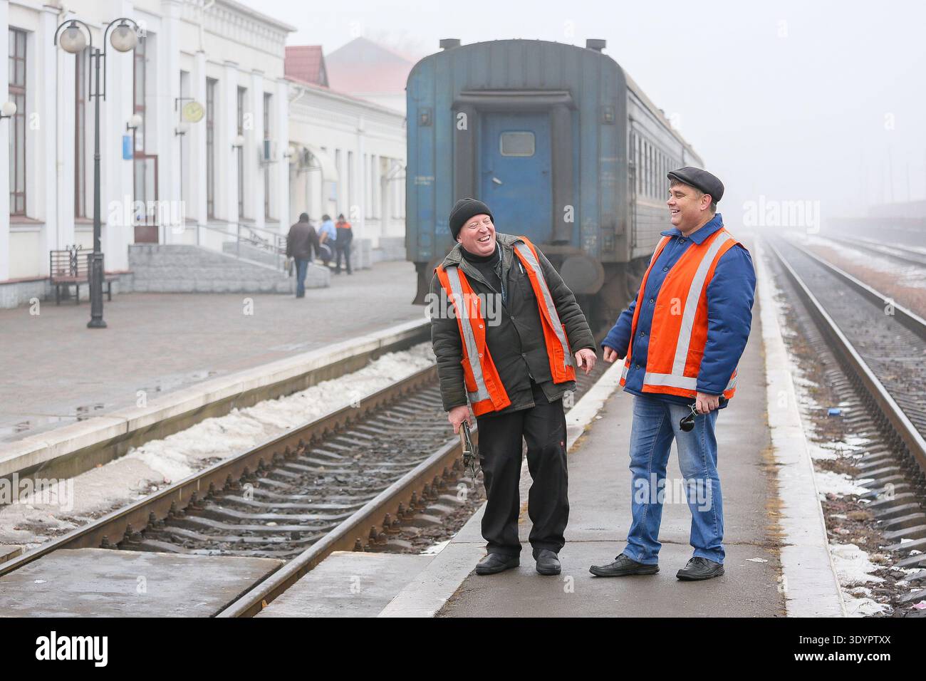January 19, 2015, Romny, Ukraine: Two railway workers in orange protective vests are chatting on a foggy platform near an old blue train Stock Photo