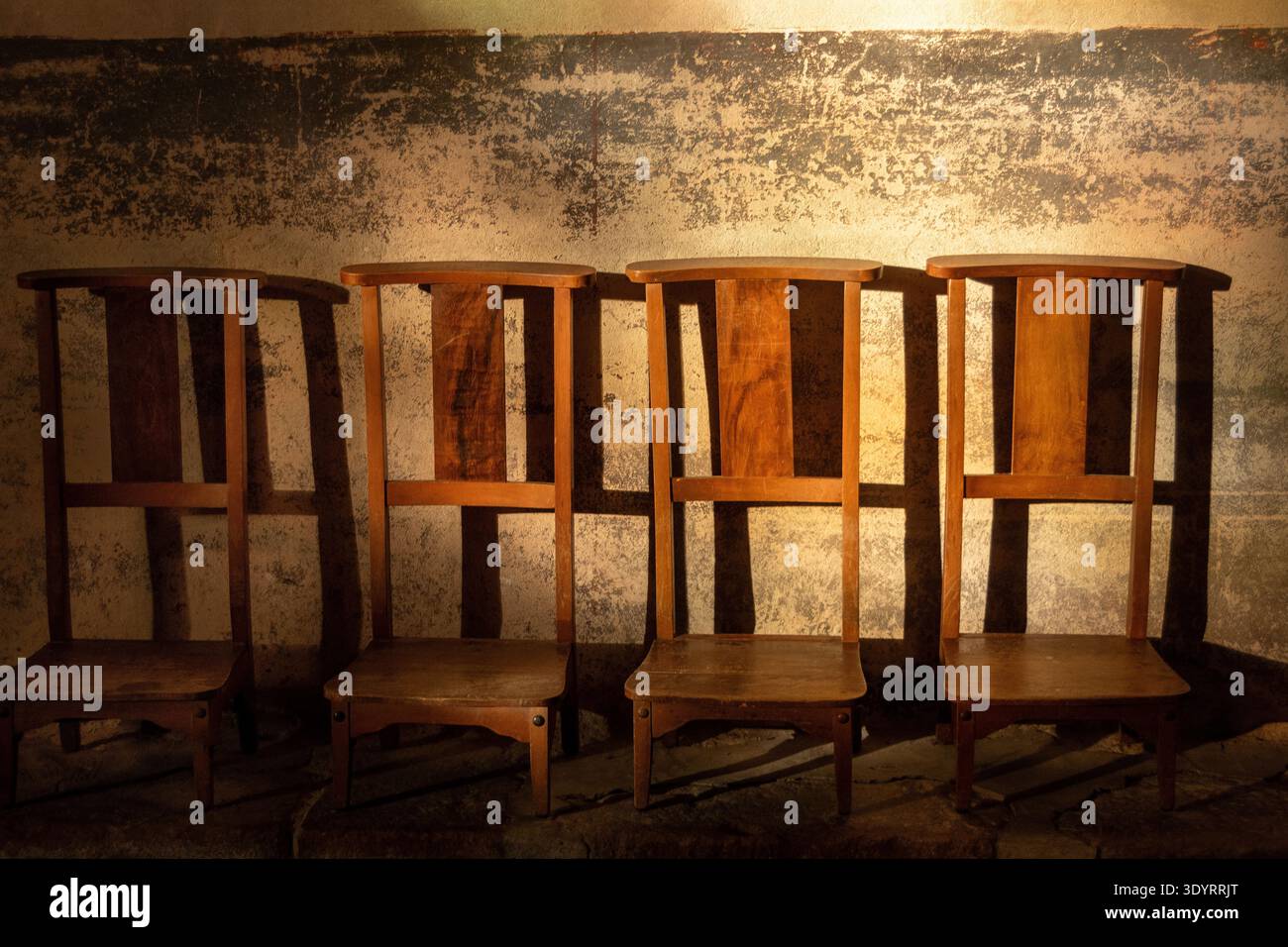 Four wooden prayer chairs sit in a row against a textured wall.The chairs are simple in design and appear aged, positioned in what looks like a church Stock Photo