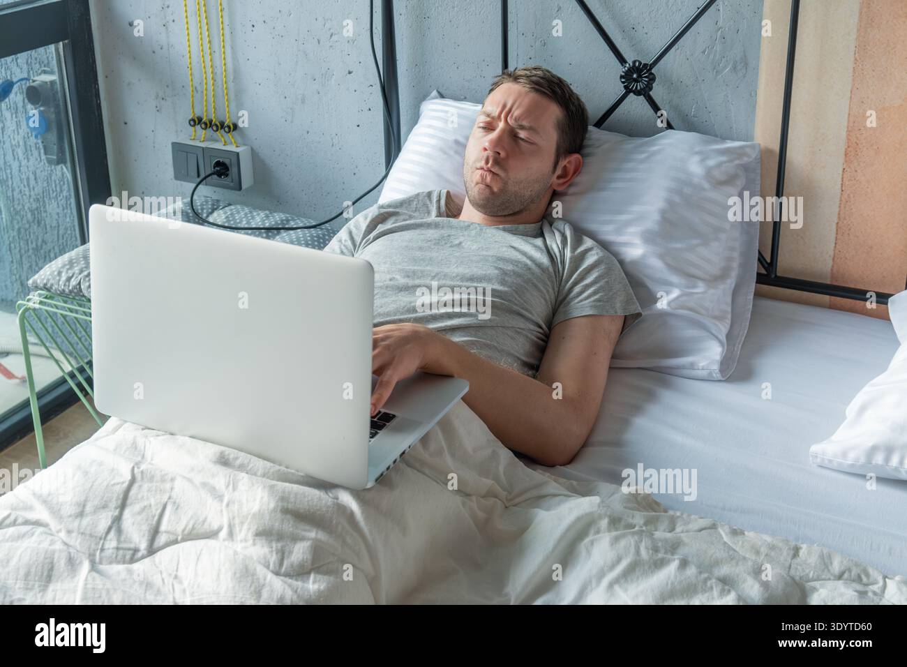 Man lying in bed working on laptop while resting with focused expression suggests concentration as he types and looks at the screen. Working from bed Stock Photo
