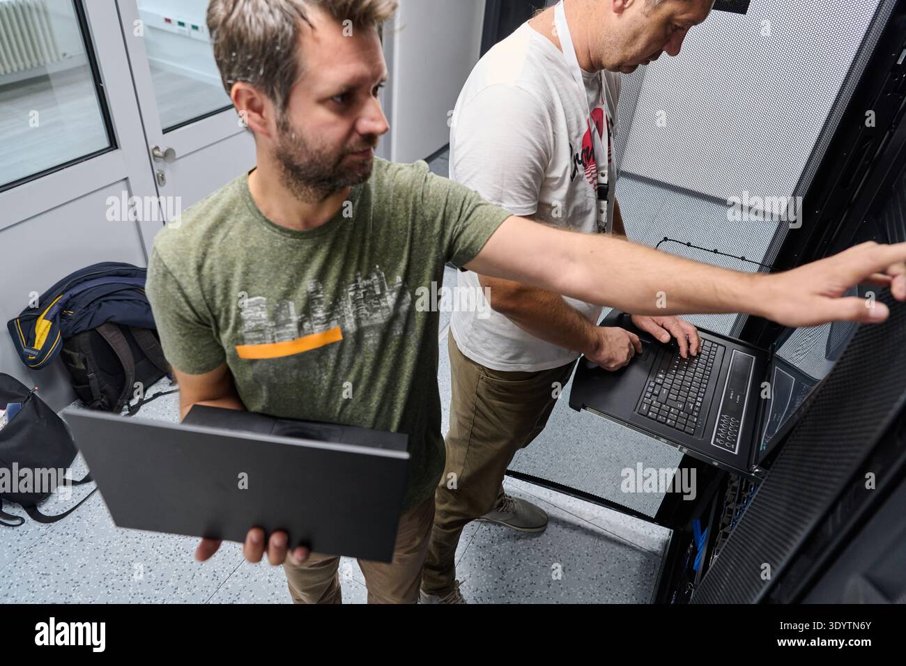 Two IT technicians troubleshooting servers in a data center rack while using laptops and collaborating Stock Photo