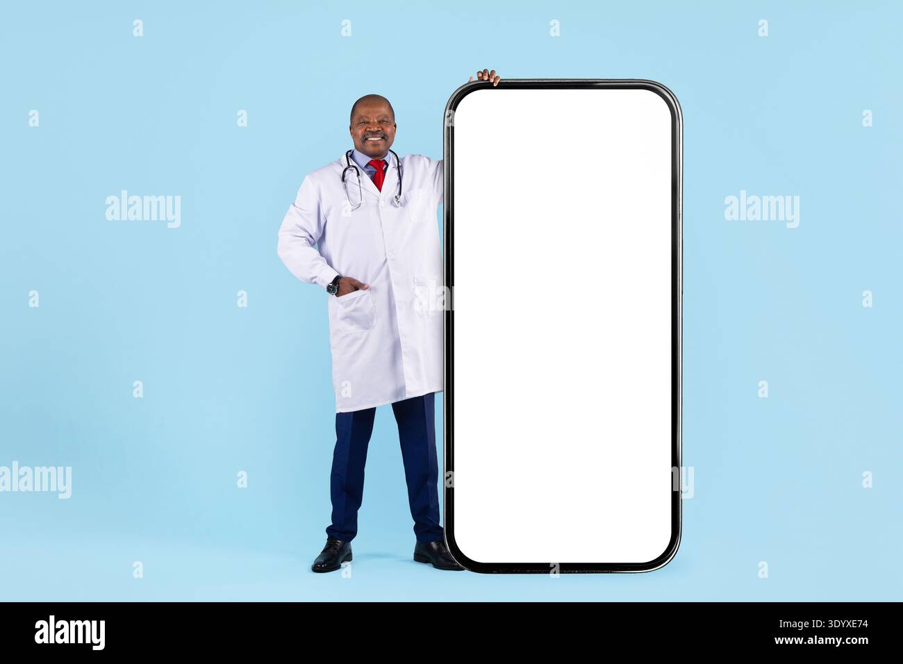 Black doctor standing next to oversized smartphone mockup on blue studio background Stock Photo