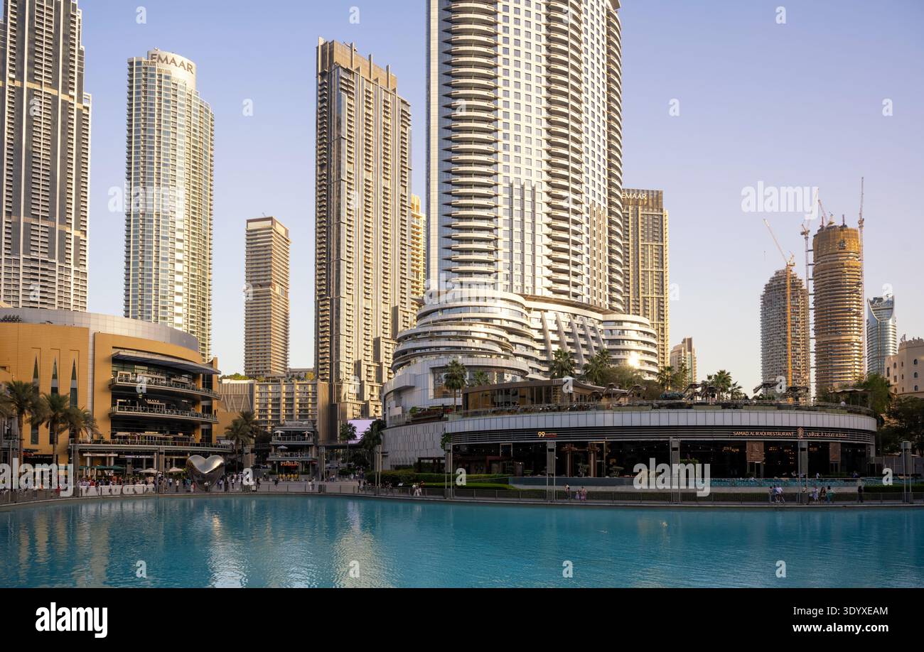A daytime exterior view of a modern downtown waterfront area. A large artificial blue lake sits in the foreground, bordered by a pedestrian promenade Stock Photo