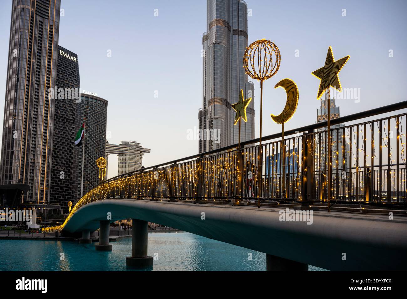 A pedestrian bridge over a body of water is adorned with warm string lights and large, illuminated golden decorations shaped like stars, a crescent mo Stock Photo