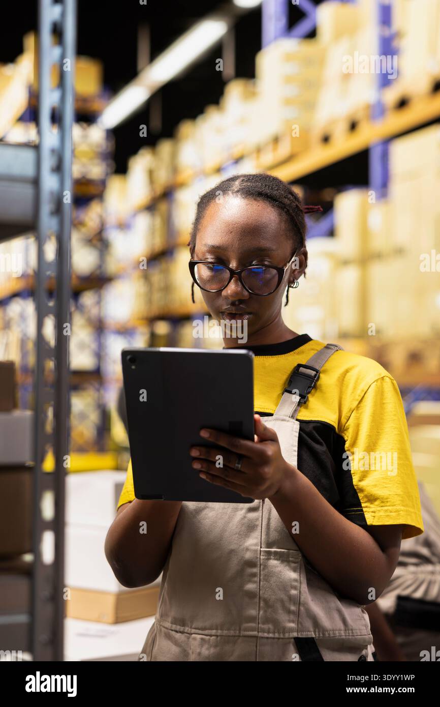 Black employee checking delivery records in warehouse logistics system on tablet. Inventory and packaging activities occur within structured shelving setup, ensuring accurate order fulfillment. Stock Photo