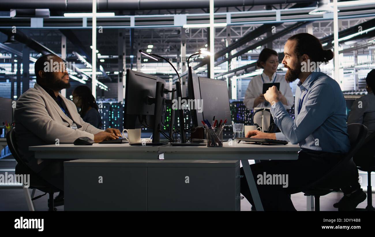 Data center technician discussing with coworker while doing AI software updates. Server room IT specialist and colleague chatting, optimizing infrastructure using artificial intelligence, camera A Stock Photo