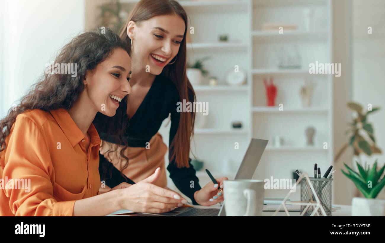 Two Women Collaborating at a Workspace While Smiling and Discussing a Project Together Stock Photo