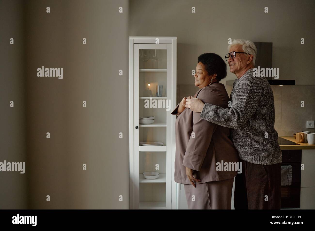 Senior Caucasian Man Embracing Senior Black Woman Standing in Kitchen Stock Photo