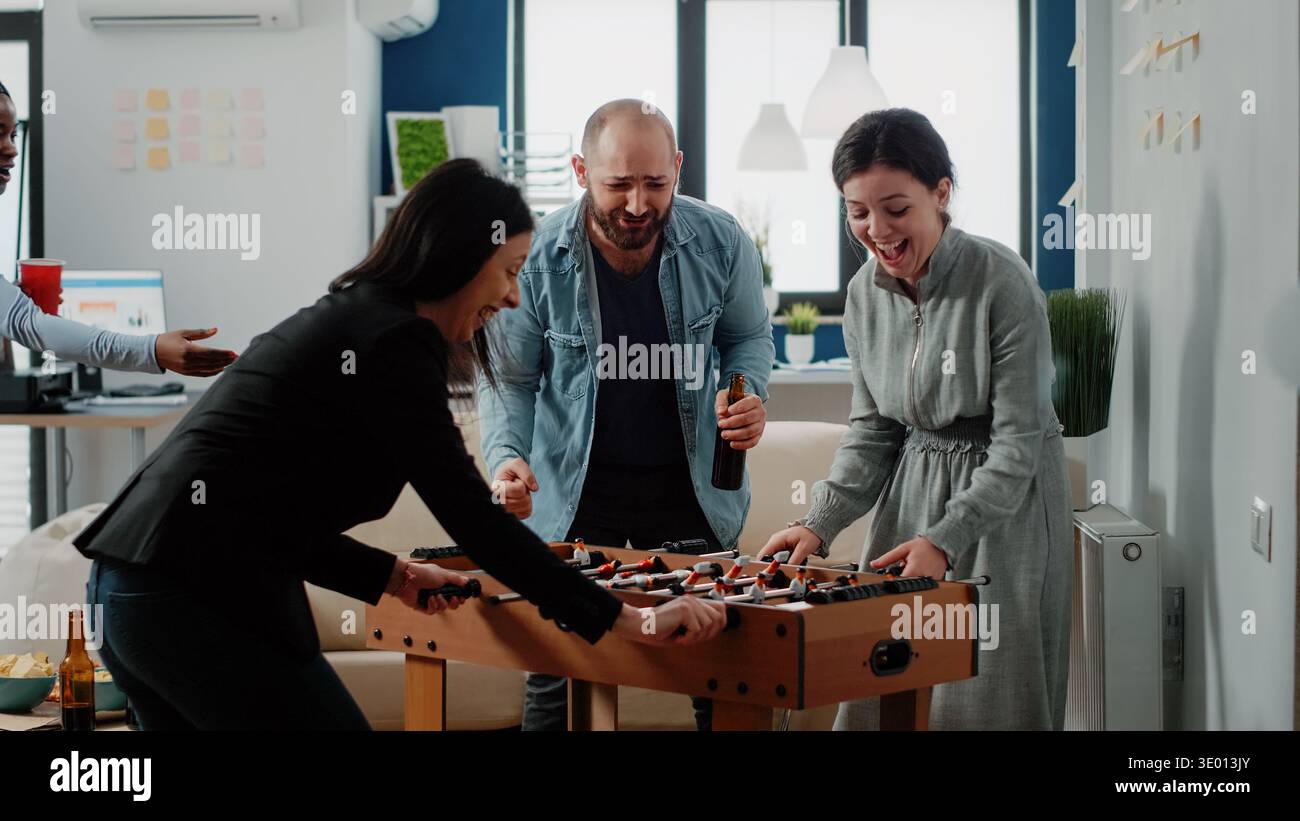 Cheerful woman dancing after winning game at foosball table at office. Coworkers playing football and enjoying fun activity after work. Multi ethnic colleagues having drinks and snacks Stock Photo