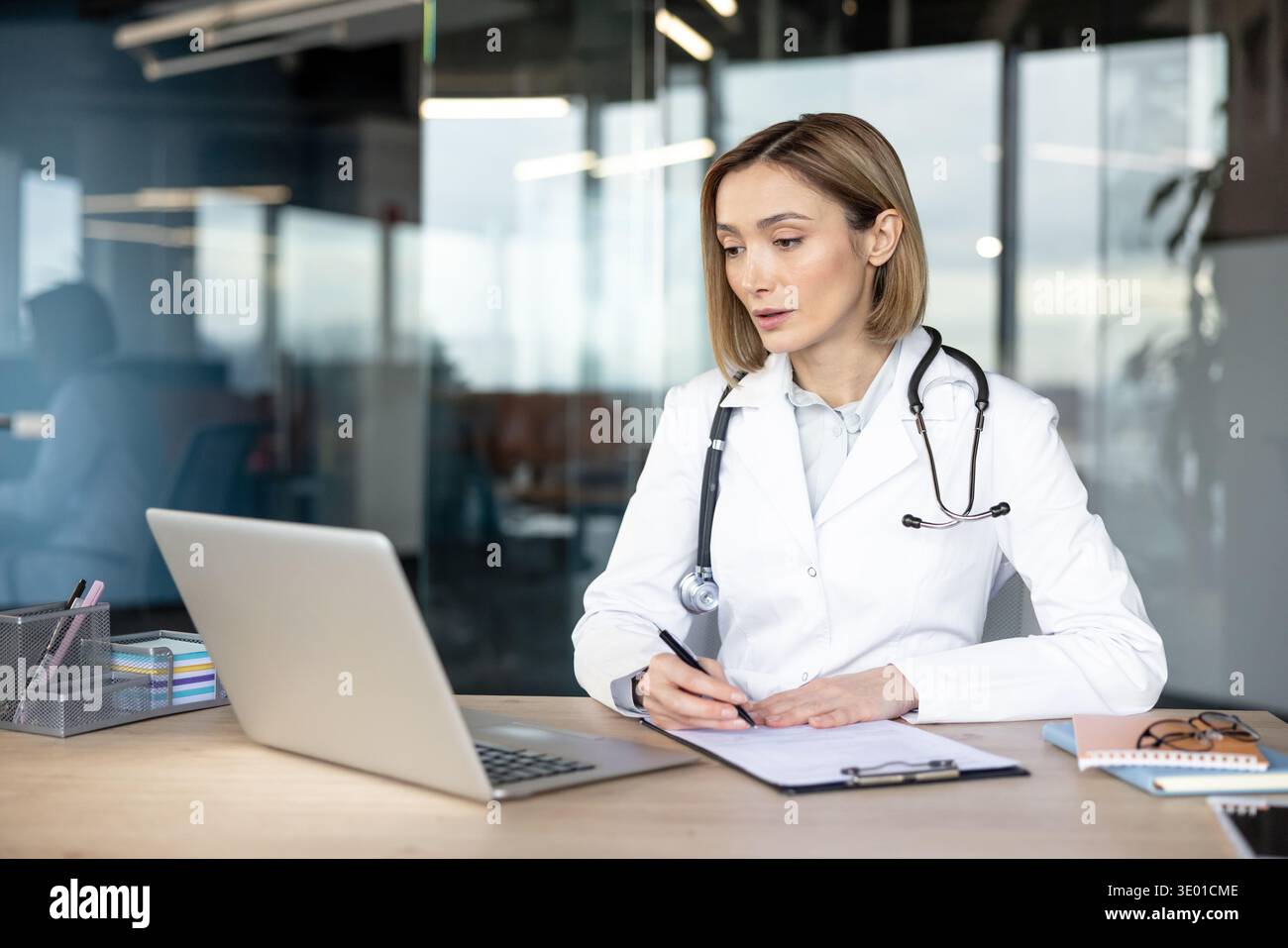 Doctor in white coat and stethoscope using laptop for medical research while writing on clipboard paper, working on healthcare documentation in a modern office Stock Photo