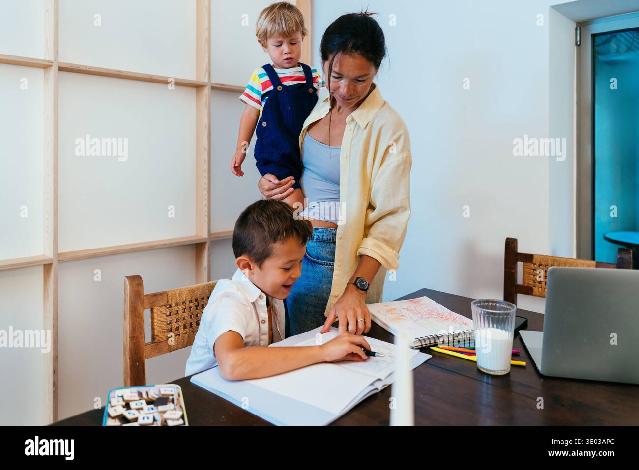 Asian family at home - asian mother helping older son with homework and drawing while holding toddler at home, supportive family learning moment Stock Photo
