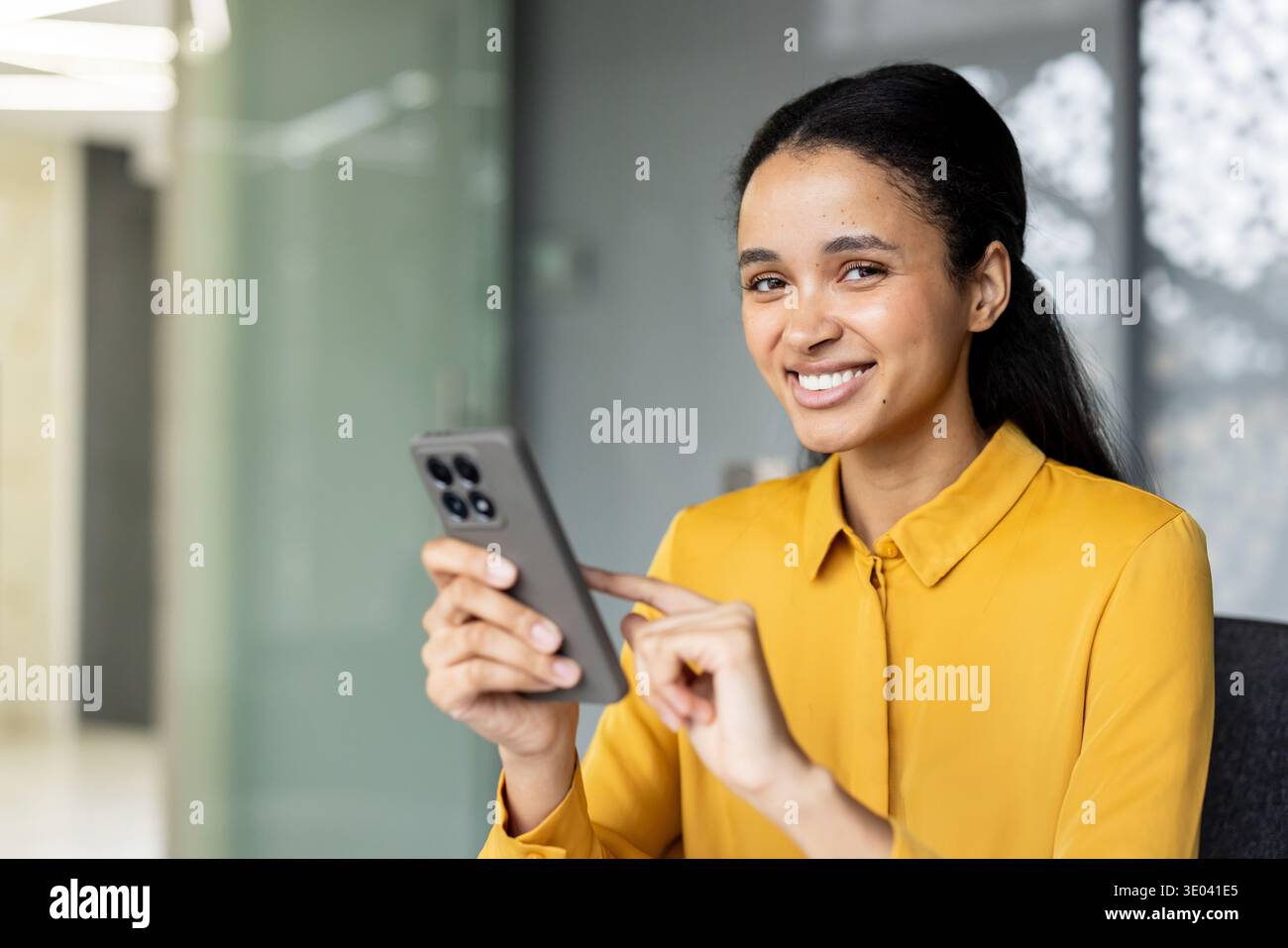 Young woman smiling at camera, holding and scrolling through a mobile phone, representing remote communication and technology in a modern office setup Stock Photo