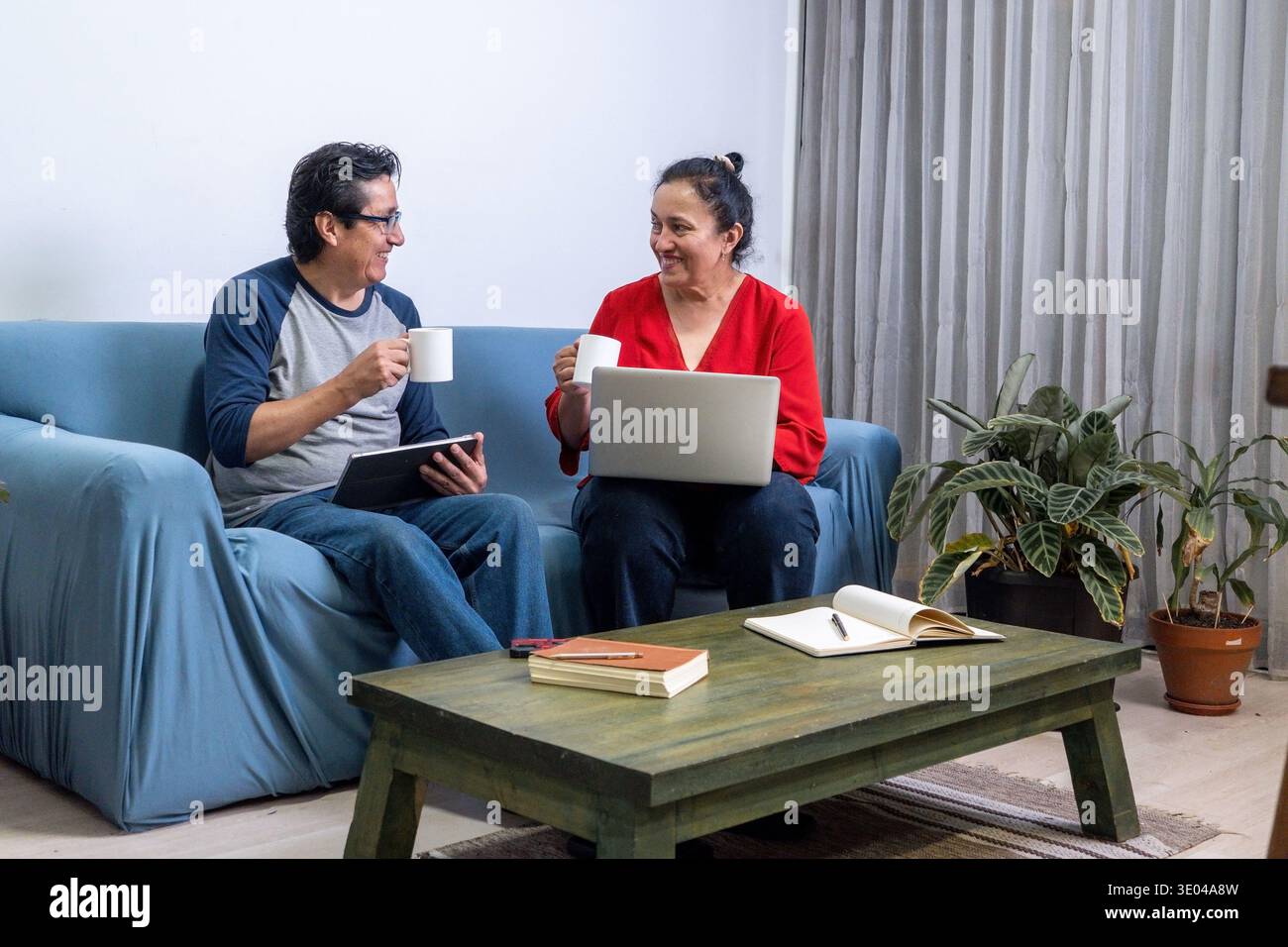Mature couple sitting on sofa using digital devices and drinking coffee together Stock Photo