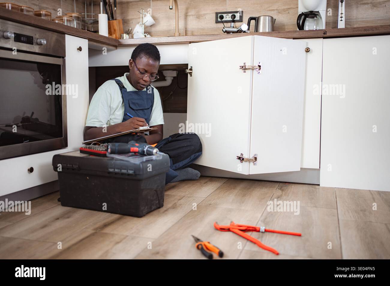 Plumber writing on clipboard while inspecting pipes under kitchen sink Stock Photo