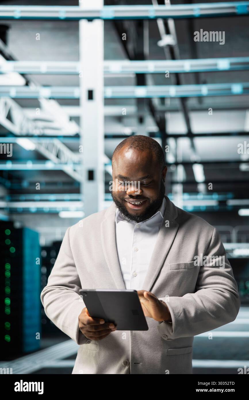 Cheerful data center technician using tablet to protect servers from exploitable software vulnerabilities, auditing systems. Jolly cybersecurity professional looking for bugs slowing down servers Stock Photo