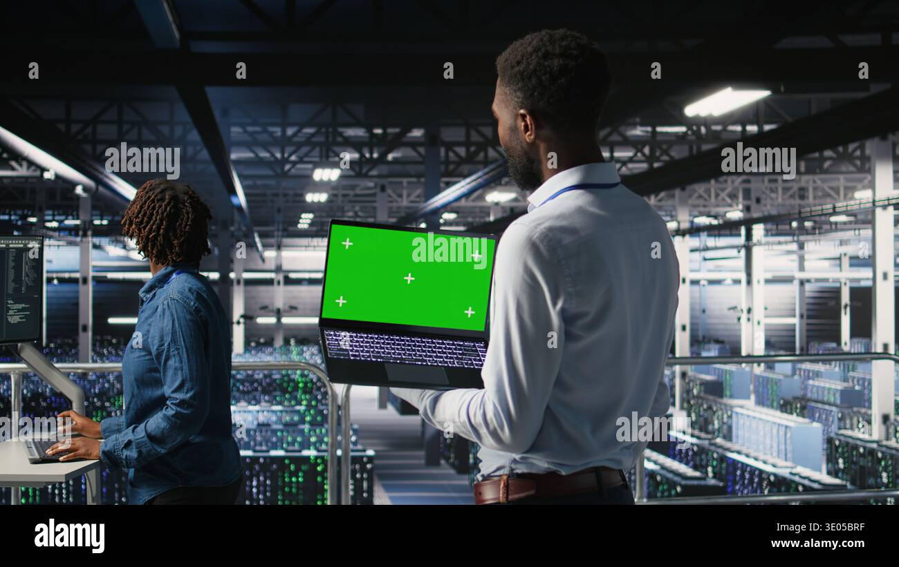 Technicians on data center industrial platform analyzing performance outputs on green screen laptop. Server hub engineering team finetuning server rig configurations using mockup notebook, Stock Photo