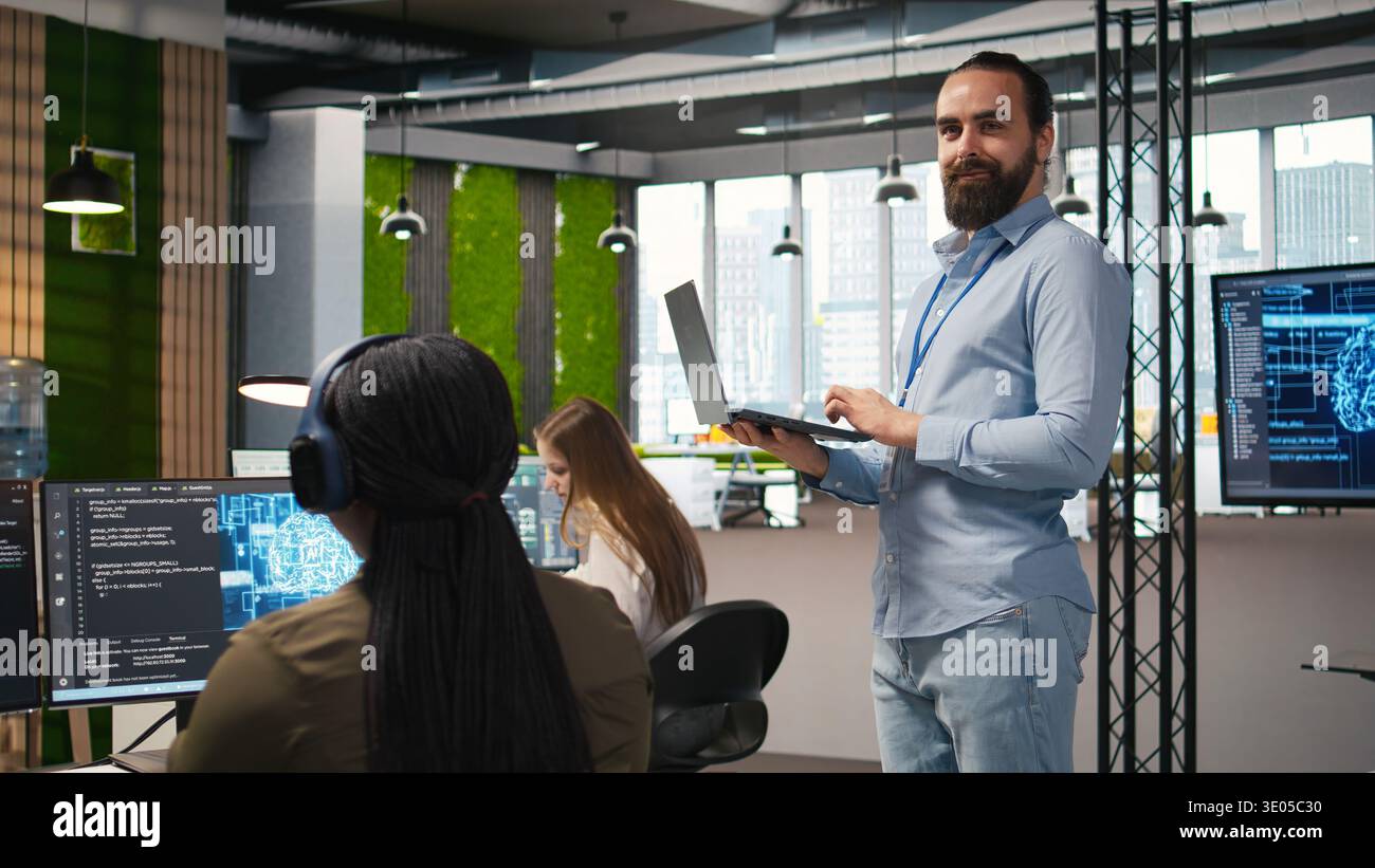 Jolly programmer coding AI algorithms on laptop to debug software issues and improving system stability. Smiling employee in tech startup office configuring settings using notebook, camera A Stock Photo