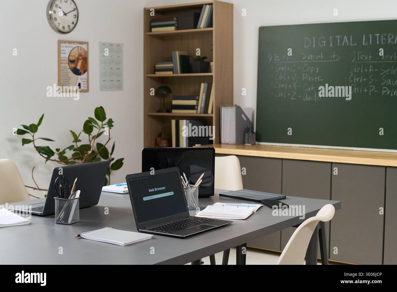 Empty classroom with several laptops on table displaying web browser error, notebooks and pens scattered around, chalkboard in background showing digital literacy course instructions Stock Photo