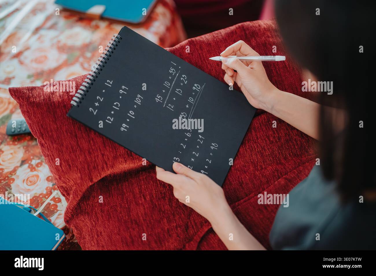 Student solving math problems in a black spiral notebook while studying at home on a red cushion Stock Photo