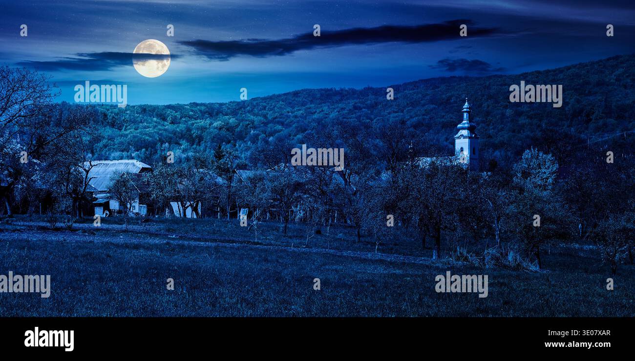 mountainous countryside landscape in spring at night. full moon over wonderful weather with cloudy sky. rural orchard in the outskirts of the village. Stock Photo