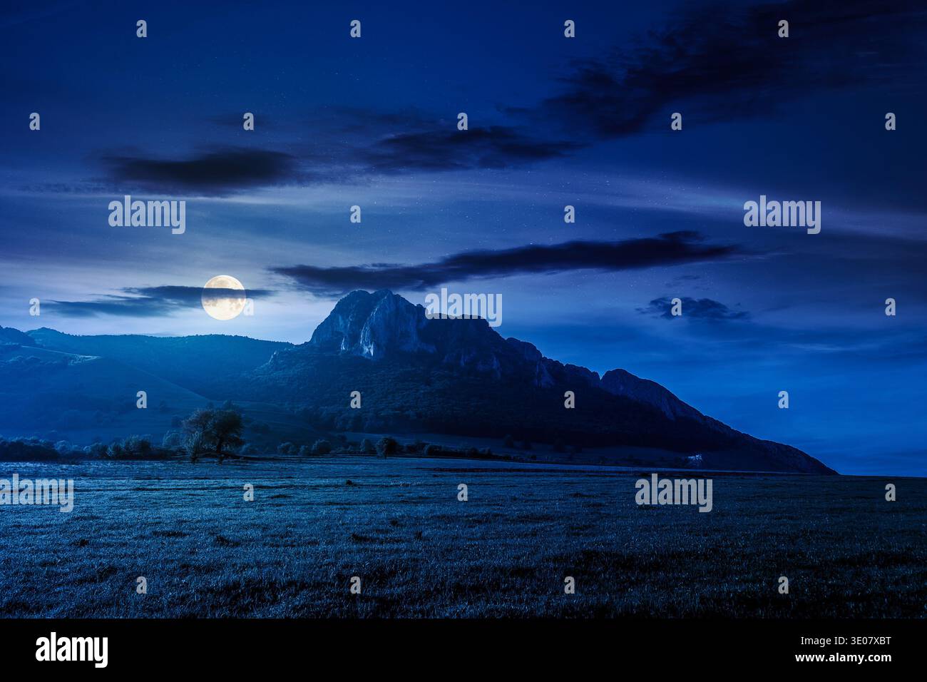 rock formation near green field at night. full moon over mysterious rural landscape in mountains. wonderful scenery lush grass in spring. clouds on th Stock Photo