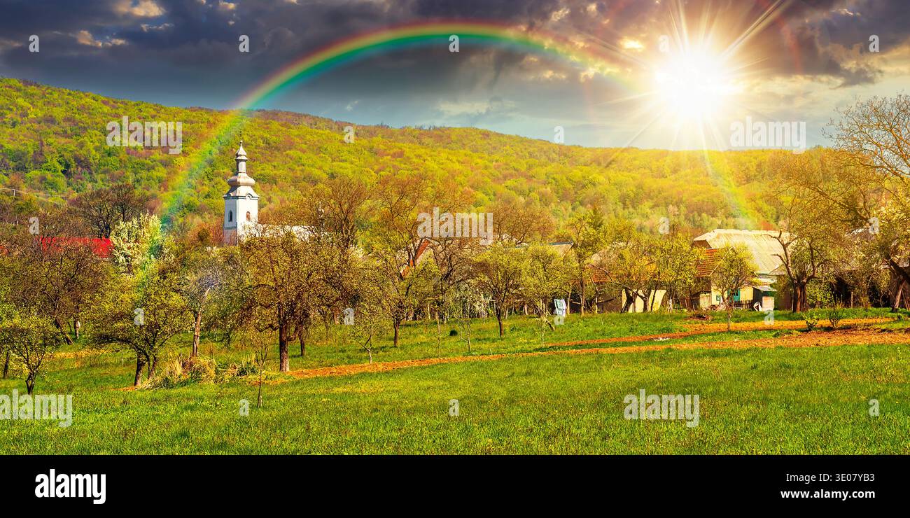 sunset over mountainous countryside landscape in spring. wonderful weather with cloudy sky and rainbow. rural orchard in the outskirts of the village. Stock Photo