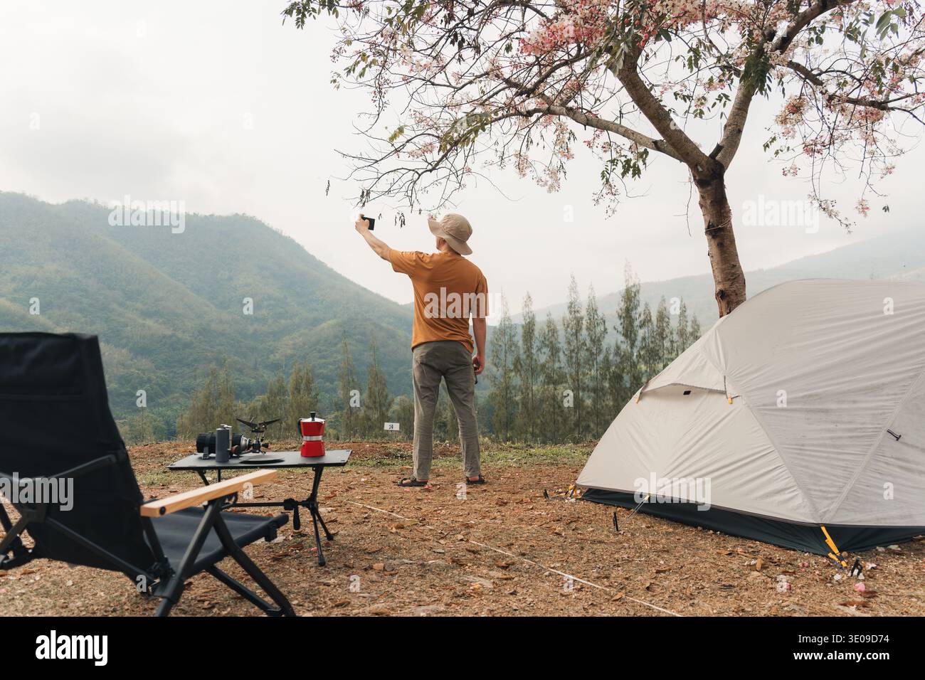 Solo traveler enjoying with using smartphone selfie at campsite with tent and camping equipment in mountain valley landscape during morning Stock Photo