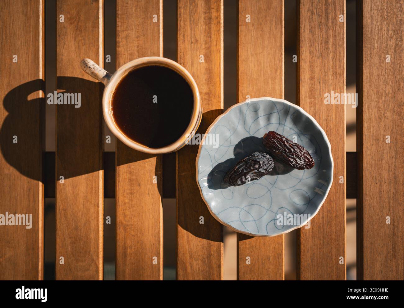 Top view of a cup of black coffee and two dried dates on a ceramic plate, arranged on a wooden table in warm morning sunlight for breakfast. Stock Photo