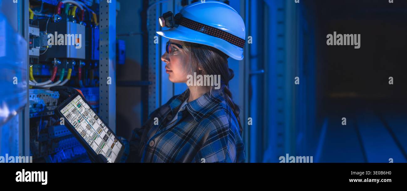 Female engineer inspecting electrical panel with tablet in dark industrial space. Engineer control. Stock Photo