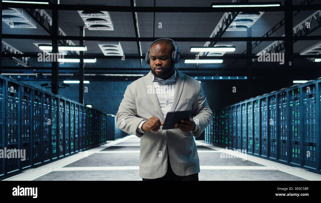 Server room programmer wearing headphones troubleshooting errors using software on tablet. IT specialist listening music, walking in data center fixing bugs using device , camera A Stock Photo