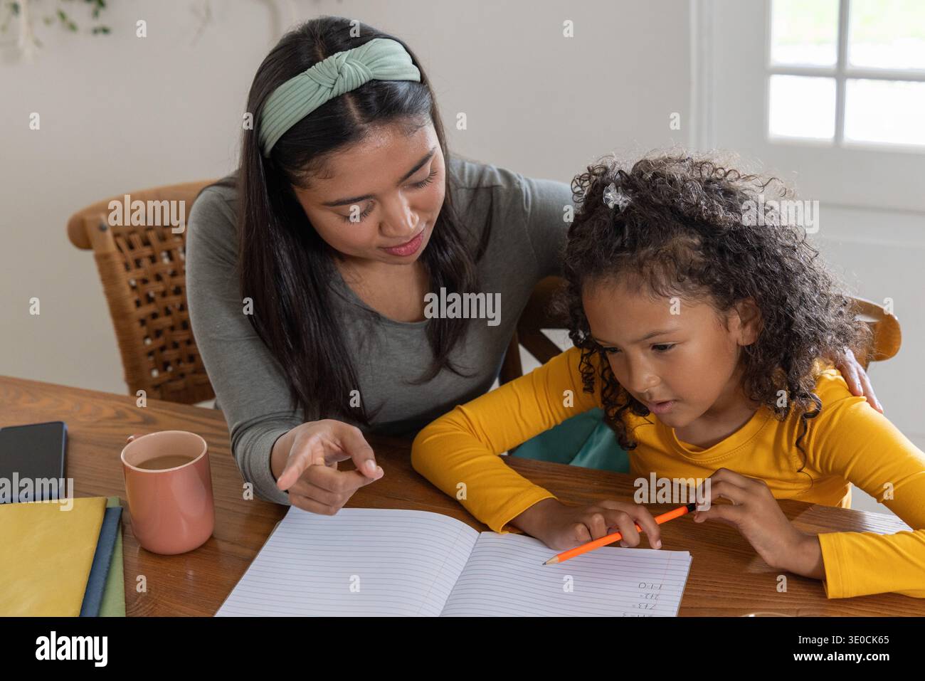 Tutor guiding child, pointing at open lined notebook on home table with orange pencil, pink mug Stock Photo
