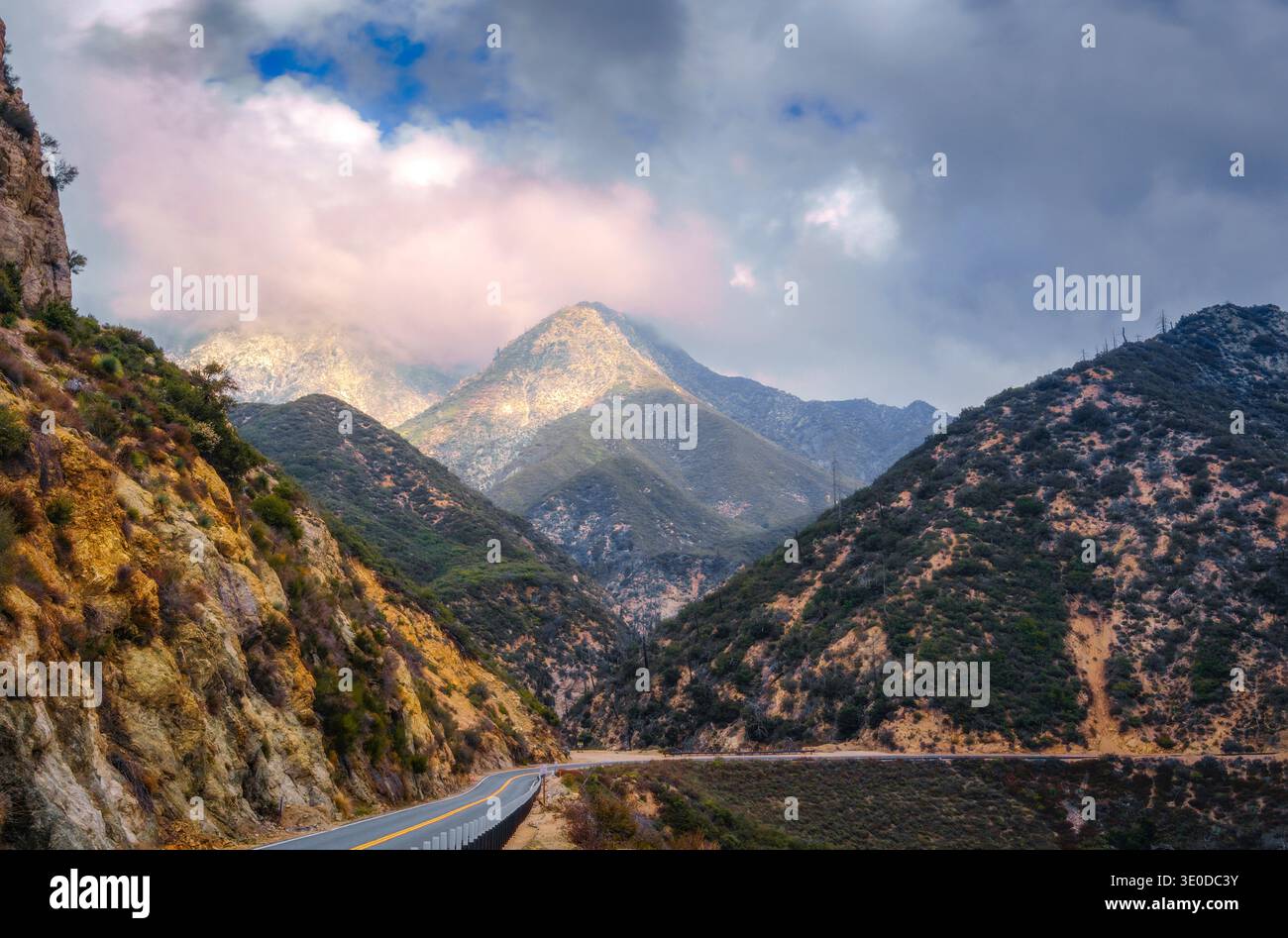 Winding mountain road passing through rugged canyon landscape beneath dramatic clouds. Stock Photo